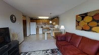 This single-floor space is designed with an open-concept layout that seamlessly connects the kitchen, dining, and living areas, creating a cohesive and functional environment. The kitchen is outfitted with warm oak cabinetry that extends to the upper walls, accented by decorative greenery along the tops of the cabinets. White appliances, including a full-size refrigerator, dishwasher, and range, are neatly integrated into the cabinetry, while a pendant light hangs above the counter, adding a touch of character. A compact dining area with a pedestal table sits at the boundary between the kitchen and living room, making it ideal for casual meals. The living area transitions smoothly from the tiled kitchen floor to plush carpeting, lending a cozy, residential feel. A patterned area rug anchors the seating zone, which features a rich red sofa and a classic floor lamp. The neutral tones of the walls and ceiling throughout the space contribute to a clean, inviting atmosphere. Overall, the layout maximizes the use of available space, offering a practical and comfortable everyday living environment.