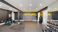 Located on Hannah Avenue in Norristown, PA, this fitness center offers a thoughtfully designed workout environment across a single floor. The main exercise area is defined by bold, dark charcoal walls accented with white trim and a striking yellow neon "stronger than yesterday" sign that serves as both motivation and a standout design element. Recessed ceiling lighting illuminates the space evenly, creating an energetic yet focused atmosphere.

The rubber-coated speckled flooring provides durability and traction throughout, making it well-suited for a range of fitness activities. Equipment includes treadmills, stationary cycles, a weight bench, a dumbbell rack, and cardio machines, offering a comprehensive workout experience. An orange upholstered bench provides a seating area, and wall-mounted mirrors and a flat-screen TV add functional detail. French doors and multiple windows along one wall allow natural light to flow in and provide views of the surrounding outdoor area.

An adjacent restroom features white painted brick walls and bold geometric patterned floor tile in black and white, adding a stylish, contemporary touch. Louvered storage closets provide practical utility within the space. The overall design balances an industrial aesthetic with modern fitness facility sensibilities, making this a well-appointed and inviting space for physical activity.