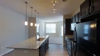 This single-floor space showcases a well-designed open-concept layout that seamlessly connects the kitchen and living area. The kitchen is a standout feature, equipped with dark espresso cabinetry that extends to the ceiling, providing ample storage. The granite countertops offer a light, speckled finish that contrasts elegantly with the dark cabinetry. A central kitchen island with an integrated stainless steel sink serves as both a functional workspace and a casual gathering point.

Lighting is a notable design element throughout the space. A row of three sleek pendant lights hangs above the island, complemented by a track lighting system on the ceiling that provides adjustable, directional illumination. The kitchen is fully equipped with black appliances, including a refrigerator and a range with overhead microwave.

The flooring transitions from hardwood-style planks in the kitchen to carpet in the adjoining living area, clearly defining each zone. The living area benefits from natural light streaming through large windows fitted with horizontal blinds. Neutral gray walls throughout create a cohesive, modern aesthetic, while standard ceiling heights contribute to a comfortable, open atmosphere.