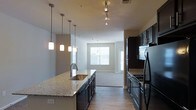 This single-floor space showcases a well-designed open-concept layout that seamlessly connects the kitchen and living area. The kitchen is a standout feature, equipped with dark espresso cabinetry that extends to the ceiling, providing ample storage. The granite countertops offer a light, speckled finish that contrasts elegantly with the dark cabinetry. A central kitchen island with an integrated stainless steel sink serves as both a functional workspace and a casual gathering point.

Lighting is a notable design element throughout the space. A row of three sleek pendant lights hangs above the island, complemented by a track lighting system on the ceiling that provides adjustable, directional illumination. The kitchen is fully equipped with black appliances, including a refrigerator and a range with overhead microwave.

The flooring transitions from hardwood-style planks in the kitchen to carpet in the adjoining living area, clearly defining each zone. The living area benefits from natural light streaming through large windows fitted with horizontal blinds. Neutral gray walls throughout create a cohesive, modern aesthetic, while standard ceiling heights contribute to a comfortable, open atmosphere.
