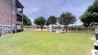 This inviting outdoor space offers a tranquil retreat defined by its direct waterfront setting and well-maintained grounds. Expansive stretches of lush, manicured lawn extend toward the water's edge, framed by a generous canopy of mature trees that provide natural shade throughout the area. At the heart of the space sits a dedicated patio area with a picnic table and a charcoal grill station, creating an ideal setup for outdoor dining and casual gatherings. The patio is positioned to maximize the sweeping views of the adjacent river, where calm waters reflect the surrounding greenery and open sky. A paved walkway traces the shoreline, offering easy access to the water's edge and encouraging leisurely strolls along the bank. The landscape is thoughtfully maintained, with mulched garden beds accenting the picnic area and adding visual interest to the grounds. In the background, residential buildings are visible, situating the space within a larger community setting. The overall ambiance is peaceful and open, characterized by abundant natural light, gentle breezes off the water, and panoramic river views that make this outdoor area a standout feature of the property.