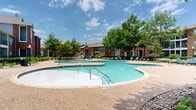 This outdoor amenity space centers around a generously sized freeform swimming pool with a smooth aggregate concrete surround that provides ample room for relaxation and recreation. The pool features a curved, resort-style shape with multiple entry points via stainless steel handrails, and depth markers are clearly visible along the edges. Surrounding the pool, a spacious sundeck is outfitted with an assortment of lounge chairs and Adirondack-style seating in blue and green, offering plenty of spots to unwind under the open sky. The area is enclosed by a black wrought iron fence with a gated entry, ensuring a secure and defined perimeter. Mature oak and deciduous trees provide generous natural shade throughout the space, complementing the well-maintained landscaping that features decorative river rock borders, lush shrubs, and trimmed hedgerows. Concrete pathway systems wind through the grounds, connecting the pool area to the surrounding multi-story brick residential buildings, which feature traditional architectural detailing including pitched rooflines and balconies. Ornamental lamp posts are positioned throughout the grounds. An adjacent brick clubhouse building with large windows is accessible directly from the pool gate, adding a functional anchor to this outdoor retreat. The overall ambiance is inviting, well-landscaped, and designed for outdoor enjoyment.