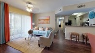 Located in the Kendall area of Miami, FL, this single-story residence offers a thoughtfully designed layout connecting living, dining, kitchen, and bedroom spaces in a cohesive flow. The living room is a welcoming focal point, showcasing dark hardwood-style flooring, a ceiling fan with light fixture, and sliding glass doors that invite natural light and provide outdoor views. The adjacent dining area features a round glass-top table beneath a pendant light, complemented by a large wall mirror that enhances the sense of space. The kitchen is well-equipped with white cabinetry, granite-style countertops, a bold stone-tile backsplash, black appliances, and matching dark wood flooring. The primary bedroom features plush carpeting, a navy and teal color palette, a large window with layered curtains, and decorative wall accents. A second bedroom carries a coastal theme, highlighted by a vivid teal accent wall adorned with tropical artwork and a bright window overlooking greenery. Two bathrooms serve the residence, each featuring granite-topped vanities with white cabinetry and dark wood-style flooring, with one offering a full tub-shower combination with white tile surround.
