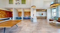 Located at 55 N Park Ave in Tucson, Arizona, this expansive community living space offers a thoughtfully designed, single-floor layout that blends contemporary style with a welcoming, social atmosphere. The space is defined by soaring double-height ceilings that amplify its generous square footage, creating an open and airy environment throughout.

The entry area makes an immediate impression with reclaimed wood accent walls, a floating built-in console, and a striking large-scale abstract painting in vibrant greens and yellows. Large-format tile flooring runs continuously throughout, lending a clean, cohesive look to the entire space. Floor-to-ceiling glass windows and a garage-style transparent door flood the interior with abundant natural light while framing panoramic views of the Tucson skyline and surrounding mountains.

The main lounge area is designed for recreation and socializing, featuring a billiards table, a ping pong table, and a shuffleboard table. A central island with bar stools provides a casual gathering spot, while a dedicated dining alcove with pendant lighting and mosaic-tiled walls offers a more intimate setting. A mounted flat-screen TV and a built-in media wall anchor a relaxed seating zone. Eclectic, artistic furnishings and curated wall art contribute to the space's vibrant, creative character, making it a dynamic hub for community life.