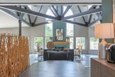 Located at 100 Runaway Bay Dr in Virginia Beach, Virginia, this single-story community clubhouse is a thoughtfully designed amenity space offering a variety of gathering and wellness areas. The centerpiece is a dramatic great room distinguished by soaring vaulted ceilings with exposed dark structural beams, rich hardwood-style flooring, and a teal-accented fireplace flanked by expansive windows that flood the space with natural light. This grand area accommodates both a comfortable lounge seating arrangement and a dining area, with direct access to the outdoors through glass doors. A sculptural driftwood room divider adds an artistic, nature-inspired touch that complements the coastal aesthetic throughout. An additional lounge nook features bold teal walls, vaulted ceilings, and curated abstract artwork, creating an intimate and stylish retreat. A dedicated conference and dining room provides a large communal table surrounded by upholstered chairs, a wall-mounted television, and a kitchenette with a refrigerator and coffee station — ideal for meetings or private gatherings. The fitness center is well-appointed with cardio equipment including treadmills, ellipticals, and a recumbent bike, as well as strength training machines and free weights, all set against deep blue accent walls with natural light from multiple windows.