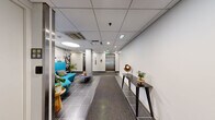 Located at 180–184 Sixth St in Cambridge, MA, this building lobby and hallway space offers a clean, professionally designed environment that serves as a functional and welcoming entry point. The layout comprises two connected hallway areas finished with large-format dark tile flooring, accented by carpet runners that guide visitors from the glass entry doors toward the elevator bank.

The elevator lobby is a central feature, housing two stainless steel elevator doors flanked by white walls with clearly marked call panels and a hand sanitizer dispenser. Overhead, a recessed drop ceiling with flush-mounted lighting and skylights provides bright, even illumination throughout the space.

A standout element is the lounge nook to one side, which features a bold teal curved sofa with accent pillows and a circular low-profile table. The nook is set against a dramatic full-scale black-and-white photographic mural depicting an industrial or rail-yard scene, adding a distinctive artistic character to the space. Potted plants and sculptural décor pieces, including a wooden sculptural form displayed on a sleek black console table, further enhance the aesthetic.

The entry corridor leading from the exterior glass doors is bright and uncluttered, offering a straightforward transition from the outdoors into the building interior. Overall, the space balances functional circulation with thoughtful design elements, creating a professional and visually engaging first impression.