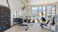 Located at 1910 S Union St in Anaheim, California, this fitness center is a thoughtfully designed, single-floor amenity space that delivers a comprehensive workout experience. The facility boasts soaring ceilings that create an open, airy atmosphere, complemented by expansive floor-to-ceiling windows that flood the interior with natural light and offer views of an outdoor pool area and the surrounding community.

The strength training zone is well-appointed with a full rack of dumbbells, adjustable weight benches, a cable machine, a multi-functional power rack with barbells, and plate-loaded selectorized machines — catering to a wide range of exercise preferences and fitness levels. The cardio section features treadmills, elliptical trainers, a stair climber, and a rowing machine, providing ample options for cardiovascular workouts.

Large wall mirrors enhance the sense of space throughout, while stylized athletic silhouette murals — depicting a BMX rider, a gymnast, a runner, and a rock climber — lend the space a dynamic, energetic character. Ceiling fans and exposed ductwork contribute to an industrial-modern aesthetic. The layout is spacious and uncluttered, allowing for comfortable movement between equipment stations and open floor training areas.