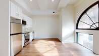 This thoughtfully designed single-floor space combines modern finishes with architectural character to create a bright and inviting environment. The open-plan living and kitchen area serves as the heart of the unit, bathed in natural light that streams through a stunning arched window — a standout architectural feature with a decorative sunburst frame that adds a classic, elegant touch. Warm hardwood flooring flows throughout, complementing the crisp white walls and coffered ceiling details that lend a sense of depth and refinement.

The kitchen is fully equipped with stainless steel appliances, including a refrigerator, range, over-the-range microwave, and dishwasher. White shaker-style cabinetry with brushed hardware extends from countertop to ceiling, maximizing storage while maintaining a clean, contemporary aesthetic. A single-basin sink and ample counter space complete the functional layout.

Additional rooms offer flexible spaces suitable for sleeping, working, or storage, each featuring the same warm hardwood floors and neutral wall tones. The bathroom is finished to a high standard, showcasing large-format marble-style floor tiles, white subway tile in the walk-in shower with a glass enclosure, and polished chrome fixtures throughout — delivering a spa-like atmosphere within a compact, well-appointed space.