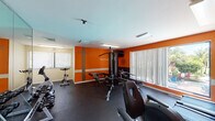 This fitness center is designed to energize and motivate, featuring bold orange accent walls adorned with inspirational words such as "Excel," "Discipline," "Conquer," "Dedication," and "Motivation." The space is laid out across a single floor and finished with durable rubber flooring throughout, providing a safe and functional workout surface. The cardio zone includes multiple treadmills, elliptical trainers, and stationary bikes from brands such as True and Sole, offering a well-rounded selection for aerobic exercise. A multi-station cable and weight machine anchors the strength training area, complemented by a rack of free weights for additional resistance training options. Full-length mirrors along one wall expand the sense of space and allow users to monitor their form. Large windows fitted with vertical blinds allow natural light to fill the room, and one window offers a direct view of an outdoor pool area with lounge seating beyond. A wall-mounted television provides entertainment during workouts. The space also includes a water cooler station stocked with sanitation supplies, ensuring a clean and comfortable environment. Overhead fluorescent lighting and ceiling-mounted security cameras complete the practical amenities of this comprehensive fitness facility.