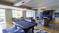 This well-appointed amenity lounge is designed to impress, offering a sophisticated blend of entertainment and relaxation within a single, thoughtfully laid-out floor. At the heart of the space sits a full-size billiards table anchored by a striking blue-and-white patterned area rug, setting the tone for the room's bold and cohesive color scheme. Large windows along one wall flood the interior with natural light, creating an open and inviting atmosphere. The space is organized into distinct functional zones: a casual seating area with plush armchairs and accent tables for conversation, a dining nook with chairs suited for more structured gatherings, and a lounge corner anchored by an elegant fireplace with a vivid cobalt blue surround. A wall-mounted flat-screen television completes the entertainment corner, making it ideal for social viewing. Pendant lighting and chandelier-style fixtures are suspended from coffered ceilings, adding architectural detail and warmth throughout. The interplay of gray walls, hardwood-style flooring, and rich blue accents gives the space a polished, upscale character that balances energy with comfort.