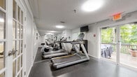 Located at 440 Meriden Rd in Waterbury, Connecticut, this single-floor exercise room offers a practical and well-appointed fitness facility. The space is finished with durable speckled epoxy flooring throughout, light gray walls, and a textured ceiling, creating a clean and functional environment.

The main workout area is equipped with cardio machines including treadmills and a recumbent bike, complemented by free weights and motivational framed artwork on the walls. Multiple windows allow natural light to filter into the fitness zone, contributing to an open and energizing atmosphere. A wall-mounted television adds convenience for users during workouts.

A set of glass-paned French doors and a sliding glass door provide direct access to an outdoor deck, where views of mature, leafy trees create a refreshing natural backdrop. This seamless indoor-outdoor connection enhances the overall ambiance of the space.

A compact kitchenette along one wall includes white cabinetry, a sink, a microwave, and a full-size refrigerator, offering added convenience. A separate area with a bistro-style high-top table and stools provides a spot to rest. Louvered closet doors offer additional storage, and safety features such as a fire extinguisher and emergency exit signage are also present throughout the space.