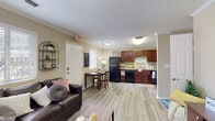 Located at 1112 E Tenth St in Greenville, NC, this single-story unit offers a well-designed, functional layout suited for everyday living. The open-concept living area features light wood-look flooring, crown molding, and large windows dressed with white blinds that invite ample natural light. The space flows seamlessly into a dedicated dining area and a fully equipped kitchen. The kitchen is appointed with rich dark wood cabinetry, granite-style countertops, and a full suite of black appliances including a refrigerator, dishwasher, oven range, and built-in microwave. A second living or flex space connects through an open doorway, offering additional room for study or relaxation. Each bedroom is comfortably sized, featuring wood-look or carpeted flooring, ceiling fans, windows for natural light, and dedicated desk areas ideal for studying. Closet storage is provided in the bedrooms. The bathrooms include both a shower stall with a glass door enclosure and a separate shower with a curtain, offering practical options for residents. Throughout the unit, warm neutral wall tones and crown molding details create a cohesive and welcoming ambiance.