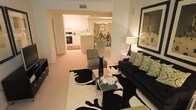 This single-floor residence offers an open-concept layout that seamlessly connects the main living areas, creating a cohesive and functional flow throughout the space. The living room serves as the heart of the home, featuring warm neutral tones, plush carpeting, and thoughtfully arranged furnishings including a dark sofa accented with green patterned throw pillows. A bold black-and-white area rug anchors the seating area, complemented by a sleek glass coffee table and decorative accents that add a polished, curated feel. A framed piece of wall art adds a finishing touch to the inviting atmosphere. Adjacent to the living room, a dedicated dining area provides an intimate setting for meals, furnished with an elegant table and chairs beneath warm ambient lighting. The kitchen is visible beyond an open passthrough, showcasing white cabinetry, a full suite of appliances including a range and dishwasher, and a breakfast bar that offers casual seating and easy access. The consistent neutral palette of warm beige tones throughout unifies the space, while recessed and accent lighting creates a welcoming ambiance. The open sightlines between rooms enhance the sense of spaciousness and connectivity across this well-designed single-level home.
