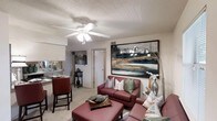 This single-floor space showcases an open-concept design that seamlessly connects the living area and kitchen. The living room serves as the focal point, furnished with a rich burgundy leather sofa and matching ottoman, accented by an assortment of decorative throw pillows in teal, coral, and green tones. A large, striking abstract canvas painting anchors the main wall, featuring deep blues, gold, and black hues that draw the eye and set a contemporary tone for the room. A ceiling fan with integrated lighting provides both airflow and illumination overhead, while a textured popcorn ceiling adds a classic touch. The kitchen is visible from the living area, separated by a raised bar counter with seating, offering a convenient casual dining option. Light-toned cabinetry and countertops in the kitchen contribute to a bright, functional workspace. Neutral carpeting flows throughout the living area, complementing the warm wall tones. Decorative floor lamps and accent vases with floral arrangements add finishing touches, enhancing the overall warmth and curated aesthetic of this well-appointed space.