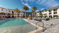 This resort-style amenity space centers around a expansive, crystal-clear pool framed by tall palm trees and a generously sized paver deck. Sun loungers, shaded cabana seating, and woven rattan pod chairs are arranged throughout the pool deck, creating numerous relaxation zones under the open sky. The surrounding multi-story residential buildings, rendered in a coastal palette of teal, sage, and white, provide an attractive architectural backdrop.

Adjacent to the pool, a single-story clubhouse lounge seamlessly blends indoor and outdoor living through large open-air openings that offer unobstructed views of the water and palm-lined landscape. Inside, the interior features smooth gray concrete flooring, exposed beam ceiling details, and abundant natural light. A central terrazzo-topped bar island with bar-height seating serves as the social hub, complemented by a bold hexagonal tile accent wall in teal, coral, and neutral tones that anchors the media display. Brass pendant lighting and wall sconces add warm, refined touches throughout.

Flexible seating arrangements—including plush sofas, rattan chairs, and dining tables—allow the space to function as a casual lounge or gathering area. The overall ambiance is bright, coastal, and inviting, with a cohesive tropical-modern aesthetic.