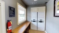 This shared laundry facility is designed with convenience and functionality in mind. The room is finished with neutral gray walls and light tile flooring, creating a clean and practical environment. At the heart of the space are two sets of stacked washer and dryer units, framed by white cabinetry with upper storage panels, providing residents with ample laundry capacity. Each unit is equipped with easy-to-use digital controls, making operation straightforward. A long wooden folding counter runs along one wall, offering generous surface space for sorting and folding laundry. The room is brightened by a large window framed in white trim, which allows natural light to fill the space and provides a pleasant view of an outdoor seating area with lounge chairs. A posted laundry center rules and guidelines sign is mounted near the entrance, keeping users informed of facility policies. Overhead, a ventilation grille ensures proper air circulation throughout the room. The overall layout is efficient and thoughtfully organized, making this laundry facility a practical and comfortable amenity for shared use.