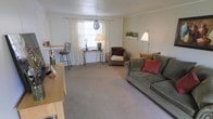 This two-floor home offers a relaxed and functional layout designed for everyday living. The living room serves as the heart of the main level, featuring wall-to-wall carpeting in a warm neutral tone that adds a sense of coziness throughout the space. A large window dressed with sheer white curtains allows natural light to filter softly into the room, creating a bright and airy atmosphere. The space accommodates a generously sized sofa accented with decorative pillows, a wicker accent chair, and a slender console table beneath the window, offering both style and practicality. A pendant light fixture hangs near the window, complementing a floor lamp positioned in the corner to provide layered lighting options. A bar-height table with stools is tucked along one wall, adding a casual dining or workspace element. Neutral-painted walls and simple crown-area trim give the room a clean, understated finish. A small shelf near the window displays subtle decorative accents, adding personality without overwhelming the space. The overall ambiance is calm and approachable, with a layout that balances open floor space with thoughtful furnishing arrangements across its two levels.