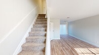 This two-story home is designed with a straightforward, functional layout that distributes living spaces across both floors. The main level features light hardwood-style flooring that flows through an open living area, which benefits from a large front-facing window that allows natural light to fill the space. A ceiling fan adds to the room's practicality. The kitchen is equipped with white shaker-style cabinetry, oil-rubbed bronze hardware, and generous granite countertops showcasing a white and grey veining pattern, providing ample prep and storage space. A staircase with carpeted treads and a clean white railing connects the two floors.

The upper level continues the neutral tone with carpeted bedrooms, each featuring windows with blinds that allow for natural light and privacy control. One room features hardwood-style flooring and a window offering views to the exterior. A full bathroom on this level includes a tub-shower combination, a toilet, and wood-style flooring, kept bright with a window and an all-white finish. Throughout both levels, neutral grey-white walls, white trim, and consistent finishes create a cohesive and move-in-ready environment.
