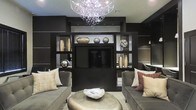 This single-floor space showcases a meticulously designed living area centered around a bold, dark aesthetic. The focal point of the room is a floor-to-ceiling built-in entertainment unit finished in deep, near-black tones, featuring open shelving that displays decorative metallic vases, sculptural accents, and a centrally mounted flat-screen television. The shelving arrangement is symmetrical, lending the space a polished, gallery-like quality.

Overhead, a striking contemporary chandelier with cascading crystal and circular glass elements draws the eye upward, adding a glamorous contrast to the otherwise dark palette. The ceiling is smooth and light-toned, providing a subtle balance against the rich, dark walls and built-ins.

Seating consists of plush, charcoal-upholstered sofas positioned facing each other, layered with cream, rust, and neutral-toned throw pillows. A low, metallic leather ottoman serves as a central coffee table, complementing the room's sophisticated material palette of dark wood, metal, and soft textiles. The overall ambiance is intimate, luxurious, and carefully curated, reflecting a modern, upscale interior design sensibility.