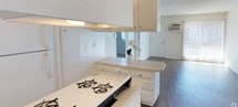 This single-floor residence offers a practical and functional layout centered around an open-concept design that seamlessly connects the kitchen and living areas. The kitchen is outfitted with white cabinetry that extends to the ceiling, maximizing storage capacity, and features a gas cooktop set into a light-toned countertop. A peninsula-style counter provides additional workspace and serves as a natural dividing point between the kitchen and the adjoining living space. The textured ceiling adds a classic residential character throughout the interior. The flooring transitions to warm wood-tone laminate or hardwood in the living area, lending a cohesive and welcoming feel to the space. The living room benefits from natural light streaming through a sliding glass door dressed with vertical blinds, which also provides direct access to the outdoors. A wall-mounted HVAC unit ensures climate control convenience. The overall palette of whites and neutral tones creates a clean, airy ambiance, making the space feel open and easy to personalize. The straightforward single-floor layout offers ease of navigation and accessibility throughout.