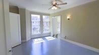 This single-floor space offers a clean, functional layout with a distinct character rooted in classic architectural details. The main room is bathed in natural light, courtesy of a set of large, multi-pane windows that overlook an urban streetscape, creating a bright and airy atmosphere throughout the day. The smooth painted floors in a cool blue-grey tone lend the space a unique, stylish finish that sets it apart from more conventional interiors. A ceiling fan with an integrated light fixture is centrally mounted, ensuring comfortable airflow and ambient lighting year-round. A wall sconce adds a warm, accent glow to complement the natural light. The entryway or adjoining area features a striking black-and-white checkered tile floor, a timeless design element that adds visual interest and a touch of classic charm. Crisp white trim and baseboards frame the neutral-toned walls throughout, contributing to a cohesive and well-maintained aesthetic. The open layout of the main area provides versatile use of the space, while the generous window placement ensures a strong connection to the surrounding urban environment. Overall, this space blends vintage character with practical design in a bright, inviting single-floor setting.