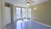 This single-floor space offers a clean, functional layout with a distinct character rooted in classic architectural details. The main room is bathed in natural light, courtesy of a set of large, multi-pane windows that overlook an urban streetscape, creating a bright and airy atmosphere throughout the day. The smooth painted floors in a cool blue-grey tone lend the space a unique, stylish finish that sets it apart from more conventional interiors. A ceiling fan with an integrated light fixture is centrally mounted, ensuring comfortable airflow and ambient lighting year-round. A wall sconce adds a warm, accent glow to complement the natural light. The entryway or adjoining area features a striking black-and-white checkered tile floor, a timeless design element that adds visual interest and a touch of classic charm. Crisp white trim and baseboards frame the neutral-toned walls throughout, contributing to a cohesive and well-maintained aesthetic. The open layout of the main area provides versatile use of the space, while the generous window placement ensures a strong connection to the surrounding urban environment. Overall, this space blends vintage character with practical design in a bright, inviting single-floor setting.