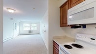 This single-floor, 2-bedroom, 1.5-bathroom residence offers a functional and well-organized layout designed for comfortable everyday living. The spacious living room features neutral carpet flooring, baseboard heating, a wall-mounted air conditioning unit, and large windows that invite natural light and outdoor views. A private entry door with a glass panel provides convenient access directly into the living area. The adjacent kitchen is well-appointed with warm medium-toned wood cabinetry, light countertops, and a full suite of white appliances including a coil-top range, built-in microwave, dishwasher, and refrigerator, all set on tile flooring. A hallway connects the living areas to the two carpeted bedrooms, each featuring natural light from double windows and baseboard heating. The primary bedroom stands out with two generous walk-in closets offering ample storage space. The full bathroom includes a tub-shower combination, a white vanity with storage, a medicine cabinet mirror, and classic white tile flooring. A separate half-bathroom provides added convenience for guests. Throughout the residence, the clean, neutral palette and straightforward design create a bright, move-in-ready living environment.