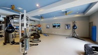 This dedicated fitness space is thoughtfully designed to accommodate a comprehensive range of workout styles and training needs. The room features a striking two-tone ceiling with recessed lighting and dual ceiling fans, creating a comfortable and well-ventilated environment for exercise. The walls are painted in a clean, neutral gray that complements the bold blue ceiling accents, lending the space a modern, energetic atmosphere.

Along one side of the room, a full-length mirror wall reflects the generous open floor area, enhancing the sense of space and allowing for form monitoring during workouts. The room is outfitted with a robust multi-functional cable and weight rack system, a battle rope, medicine balls, stability balls, and a balance trainer, providing everything needed for strength, cardio, and functional training. A standing heavy bag and its support rack add a boxing and martial arts dimension to the fitness offering.

Three wall-mounted flat-screen TVs are positioned for easy viewing from various points in the room, ensuring an engaging workout experience. Framed artwork adds a polished, finished look to the space. The light-colored flooring provides a clean, open workout surface. Overall, this fitness room combines practicality with a professional aesthetic, making it a fully equipped destination for serious training.