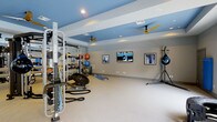 This dedicated fitness space is thoughtfully designed to accommodate a comprehensive range of workout styles and training needs. The room features a striking two-tone ceiling with recessed lighting and dual ceiling fans, creating a comfortable and well-ventilated environment for exercise. The walls are painted in a clean, neutral gray that complements the bold blue ceiling accents, lending the space a modern, energetic atmosphere.

Along one side of the room, a full-length mirror wall reflects the generous open floor area, enhancing the sense of space and allowing for form monitoring during workouts. The room is outfitted with a robust multi-functional cable and weight rack system, a battle rope, medicine balls, stability balls, and a balance trainer, providing everything needed for strength, cardio, and functional training. A standing heavy bag and its support rack add a boxing and martial arts dimension to the fitness offering.

Three wall-mounted flat-screen TVs are positioned for easy viewing from various points in the room, ensuring an engaging workout experience. Framed artwork adds a polished, finished look to the space. The light-colored flooring provides a clean, open workout surface. Overall, this fitness room combines practicality with a professional aesthetic, making it a fully equipped destination for serious training.