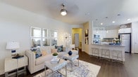 This single-floor residence showcases an open-concept design that integrates the living and kitchen areas into one cohesive, functional space. The living area is anchored by a neutral-toned sofa adorned with blue and patterned accent pillows, complemented by a mirrored wall arrangement that enhances the sense of depth and light. A decorative starburst floor lamp and a ceiling fan with a polished chrome finish add character to the space. Dark hardwood flooring runs throughout, providing a warm contrast to the light-colored walls and furnishings. A patterned area rug in blue and cream tones defines the seating area, which also features a sleek glass-and-metal coffee table.

The kitchen is positioned adjacent to the living space and features white cabinetry, granite or stone countertops, stainless steel appliances, and a peninsula with bar-height seating. Pendant lighting with a chandelier-style fixture illuminates the kitchen, adding an elegant touch. A hallway visible in the background leads to additional areas of the residence. Overall, the space exudes a bright, contemporary ambiance with a cohesive blue-and-white color palette.