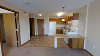 This single-floor unit offers a practical and well-organized open-concept layout that seamlessly connects the kitchen and living areas. The kitchen is outfitted with warm honey-toned wood cabinetry providing ample storage, a white refrigerator, an electric range, and a sink positioned beneath upper cabinets. A peninsula with a light-toned countertop extends into the space, offering a convenient breakfast bar and additional prep area. Pendant lighting hangs above the peninsula, adding a focused, inviting glow to the cooking and dining zone, while flush-mount ceiling fixtures illuminate the broader space. The flooring transitions from tile in the kitchen area to plush carpet in the living space, visually delineating the two zones while maintaining an open feel. Neutral-toned walls throughout give the space a clean, versatile backdrop. A hallway leads to what appears to be additional rooms, including what is visible as a closet or utility door, suggesting a functional layout designed to maximize usability. The overall ambiance is cozy and practical, suited for everyday living with an emphasis on accessibility and ease of movement throughout.