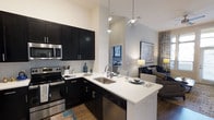 This single-floor residence showcases a thoughtfully designed open-concept layout that seamlessly connects the kitchen and living areas. The kitchen is a standout feature, boasting dramatic vaulted ceilings that amplify the sense of space and airiness. Dark espresso cabinetry provides bold contrast against crisp white quartz countertops, while a full suite of stainless steel appliances — including a range, microwave, and dishwasher — adds a sleek, modern finish. A subway tile backsplash complements the overall aesthetic. The peninsula island with an undermount double sink serves as both a functional workspace and a casual dining or entertaining surface, anchored by stylish pendant lighting overhead. Wood-style flooring runs throughout, adding warmth to the contemporary palette. The adjacent living area is bright and welcoming, benefiting from the open sightlines and natural light filtering in from nearby windows. The neutral wall tones and high ceilings contribute to a clean, airy ambiance, making the space feel expansive and well-appointed throughout.