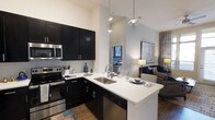 This single-floor residence showcases a thoughtfully designed open-concept layout that seamlessly connects the kitchen and living areas. The kitchen is a standout feature, boasting dramatic vaulted ceilings that amplify the sense of space and airiness. Dark espresso cabinetry provides bold contrast against crisp white quartz countertops, while a full suite of stainless steel appliances — including a range, microwave, and dishwasher — adds a sleek, modern finish. A subway tile backsplash complements the overall aesthetic. The peninsula island with an undermount double sink serves as both a functional workspace and a casual dining or entertaining surface, anchored by stylish pendant lighting overhead. Wood-style flooring runs throughout, adding warmth to the contemporary palette. The adjacent living area is bright and welcoming, benefiting from the open sightlines and natural light filtering in from nearby windows. The neutral wall tones and high ceilings contribute to a clean, airy ambiance, making the space feel expansive and well-appointed throughout.