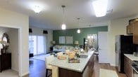 Located at 740 Bridgeport Avenue in Shelton, Connecticut, this single-floor residence offers a thoughtfully designed open-concept layout that promotes a sense of spaciousness and effortless flow. The kitchen serves as the heart of the home, featuring a large granite-topped island with bar seating, dark wood cabinetry, a stainless steel sink, and elegant pendant lighting that adds a contemporary touch. The kitchen transitions seamlessly into the living area, which is anchored by a bold, deep green accent wall that brings warmth and character to the space. Rich hardwood flooring runs throughout, tying the rooms together cohesively. The living area is well-lit, benefiting from natural light streaming in through sliding glass doors that open to the outdoors, enhancing the bright and airy ambiance. Recessed and overhead lighting ensure the space is well-illuminated at all hours. The overall design aesthetic balances modern finishes with inviting, comfortable elements, making it ideal for both entertaining and everyday living. The combination of stylish décor, quality materials, and an intelligently planned layout makes this a highly functional and visually appealing residence.
