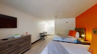 This 2-Bed Queen Studio in Memphis, Tennessee, is a thoughtfully laid-out single-floor unit designed to accommodate two guests comfortably. The main living area features two queen-sized beds, each outfitted with clean white linens and accented with vibrant orange pillows that complement the bold orange feature wall — a defining design element that adds energy and personality to the space. Rustic wood-framed headboards and matching wood-toned dressers provide practical storage while contributing a cohesive, modern aesthetic. Light wood-style flooring runs throughout, lending warmth and easy maintenance to the unit.

A compact dining area with a small table and two chairs sits centrally within the studio, offering a convenient space for meals. Adjacent to the dining area, a well-equipped kitchenette provides essential appliances including a refrigerator, microwave, stovetop, and ample white cabinetry for storage — ideal for self-sufficient stays.

The bathroom is clean and functional, featuring a shower with a bold orange-and-white striped curtain, a wall-mounted towel rack stocked with fresh linens, a vanity counter, and a toilet. A window with vertical blinds allows natural light into the sleeping area, while a wall-mounted HVAC unit ensures year-round climate control. The textured ceiling and neutral walls round out this practical, inviting studio.