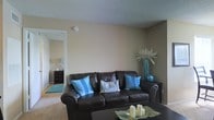 This single-story space centers around a welcoming living area designed with a clean, functional aesthetic. The room features neutral beige walls paired with a textured ceiling, creating a warm and understated backdrop. A large, dark brown leather sofa serves as the focal point, dressed with vibrant teal and patterned throw pillows that introduce a pop of color and visual interest to the space.

A sleek, dark coffee table sits in front of the sofa, accented with decorative candles and woven placemats, adding a layered, curated touch. To the side, a tall decorative vase with an ornamental branch arrangement complements the teal color scheme and adds vertical dimension to the room.

The flooring appears to be a light-toned carpet, contributing to the room's comfortable feel. A doorway visible from the living area leads to an adjacent room, which features a dark wood dresser and area rug, suggesting a bedroom or secondary living space. The overall ambiance is casual yet thoughtfully arranged, making the space feel inviting and livable, with cohesive décor details tying the areas together.