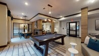This single-floor amenity lounge is thoughtfully designed to offer residents a variety of social and recreational spaces within one cohesive layout. The space is anchored by a central billiards area, where a full-size pool table rests atop a geometric area rug beneath a statement pendant light fixture, creating a natural gathering point for entertainment.

Adjacent to the billiards zone is a comfortable lounge area furnished with plush neutral sofas, teal accent chairs, and a rustic wood coffee table. A paneled accent wall provides a refined architectural backdrop, and curated artwork adds a polished, contemporary touch. Recessed and pendant lighting throughout ensures a warm, inviting ambiance.

A built-in wet bar and media nook offer additional entertainment functionality, featuring dual flat-screen televisions, wood cabinetry, and pendant lighting that complements the teal upholstered seating arranged in front.

The private dining area accommodates a large group around a solid wood table surrounded by modern teal chairs. Floor-to-ceiling glass doors open directly to an outdoor terrace equipped with grilling stations, seamlessly extending the entertaining space outdoors.

The entry corridor features large-format tile flooring, glass-paneled doors, and key-access security, providing a clean and secure transition into the lounge. Overall, the space balances modern design with functional amenities throughout.