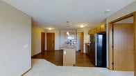 This single-floor space showcases an open-concept layout that seamlessly connects the living area to the kitchen, creating a functional and cohesive environment. The kitchen is well-appointed with stainless steel appliances, including a refrigerator and electric range, complemented by warm honey-toned wood cabinetry that extends to the upper walls for ample storage. A center island with a light wood finish serves as both a prep space and a casual gathering point, enhanced by pendant lighting overhead that adds a focused, warm glow to the area. The floors transition from durable hardwood-style flooring in the kitchen and entry zones to plush carpeting in the living area, defining each space naturally. Flush-mounted ceiling lights provide consistent illumination throughout. A solid wood entry door with a deadbolt is visible, offering a sense of security and a welcoming focal point. Additional doors lead to what appear to be a closet and a secondary room, suggesting practical storage and functional room separation. The overall ambiance is warm and understated, with neutral walls and quality finishes that provide a versatile foundation for personalization.