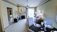This thoughtfully designed single-floor residence offers a seamless blend of comfort and refined style. The living area serves as the heart of the space, anchored by a classic arched fireplace with a decorative mantle adorned with curated accents, including a round sunburst mirror and artistic wall décor. Soft, neutral tones on the walls complement the plush carpeting, creating a cohesive and calming palette throughout.

A ceiling fan with a brushed nickel finish is centrally positioned, adding both functionality and a subtle design element to the room. Large arched windows dressed with layered drapes allow generous amounts of natural light to pour in, enhancing the open and airy feel of the space. The seating arrangement features a plush sofa with coordinating throw pillows, flanked by warm-toned wooden side tables and elegant table lamps that add a cozy glow.

The dining area is defined by a sleek dark round table set with coordinating upholstered chairs, positioned to take advantage of the living area's inviting ambiance. A dark wood media console with cabinet storage adds practical functionality along one wall. Together, these elements create a cohesive living environment that balances everyday comfort with an elevated, well-appointed aesthetic.