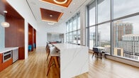 This contemporary Texas A&M University amenity floor is a thoughtfully designed space that blends academic identity with upscale, modern interiors. The open-plan layout is anchored by dramatic floor-to-ceiling glass curtain walls that frame sweeping panoramic views of the surrounding urban skyline, flooding the interior with abundant natural light throughout the day.

A striking white marble-veined island with warm wood bar stools serves as the centerpiece of the main gathering area, complemented by a distinctive recessed copper-toned ceiling feature with circular pendant lighting above. Light hardwood flooring runs throughout, lending warmth and continuity across the space.

Multiple seating zones cater to different needs: casual lounge areas feature plush mint-green upholstered chairs arranged around a patterned area rug, while a vibrant multicolored striped banquette anchors a social dining nook near a kitchenette finished with seafoam tile, pendant globe lights, and built-in appliances. A rich wood-paneled wall houses a large mounted display screen, creating a dedicated media and relaxation area.

Glass-enclosed conference and meeting rooms offer more private settings without sacrificing the connection to natural light and views. The overall ambiance is energetic yet refined, reflecting a space designed to inspire collaboration, connection, and comfort.