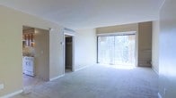 This two-floor residence offers a straightforward and functional layout designed for comfortable living. The main living area is finished with wall-to-wall carpeting in a neutral tone, complementing the warm beige walls that create a clean, understated aesthetic throughout the space.

A prominent sliding glass door, dressed with full-length vertical blinds, serves as a central feature of the living area. It provides an abundance of natural light and offers views of the surrounding greenery, creating a bright and pleasant atmosphere. The door likely leads to an outdoor space such as a patio or balcony.

A walk-in closet is conveniently accessible from the living area, providing generous storage capacity. The closet is fitted with a hanging rod and open shelving, maximizing its organizational potential. An overhead HVAC vent is positioned above the closet entrance, ensuring consistent climate control throughout the room.

The transition between flooring materials — carpet in the living area shifting to tile near the entryway — adds subtle definition between functional zones. Spread across two floors, the residence provides versatile living arrangements suitable for a variety of needs.