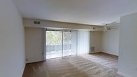 This single-floor residence offers a clean, functional layout designed for comfortable everyday living. The main living area is the focal point of the space, characterized by its open feel and an abundance of natural light streaming in through large sliding glass doors. The doors are dressed with vertical blinds that allow for flexible light control and privacy, while offering a view of a balcony and the lush greenery beyond. A ceiling fan is centrally positioned to promote airflow and comfort throughout the room. The interior features neutral-toned walls and plush wall-to-wall carpeting that adds warmth underfoot. Practical HVAC vents are integrated into both the ceiling and the lower wall, ensuring year-round climate comfort. The overall aesthetic is straightforward and move-in ready, with a light, airy ambiance that makes the living space feel welcoming and versatile. The seamless connection between the interior and the outdoor balcony area enhances the sense of openness, making this space well-suited for both relaxed daily living and casual entertaining.