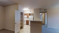 This single-floor residential unit offers a practical and well-organized layout designed for comfortable everyday living. The kitchen serves as a central feature of the home, equipped with white appliances including a refrigerator, electric range, and microwave. Warm-toned wooden cabinetry provides ample storage, while a built-in pantry with open shelving adds additional organization near the kitchen entrance. A breakfast bar with a light-toned countertop extends from the kitchen, creating a natural transition into the adjoining living area and offering a convenient space for casual dining or entertaining. The flooring transitions from light tile in the kitchen to carpeting in the living and hallway areas, adding warmth and definition to each zone. Neutral beige walls and standard overhead lighting give the space a clean, understated aesthetic. A hallway visible from the main living area leads toward additional rooms, suggesting a thoughtful floor plan that balances open communal spaces with private areas. HVAC vents are present throughout, ensuring climate comfort. Overall, the unit presents a functional and move-in-ready environment suited for a range of lifestyles.