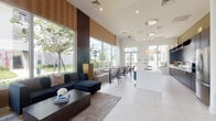 Located at 7065 Banyan Leaf Dr in West Palm Beach, FL, this single-story community clubhouse offers a sophisticated and welcoming environment designed with modern Florida living in mind. The space is defined by its soaring ceilings and expansive floor-to-ceiling windows that flood the interior with natural light and frame lush outdoor greenery and palm trees beyond.

The grand entrance foyer sets an elegant tone, featuring large-format white marble-look tile flooring, double glass entry doors with transom windows, and tasteful accent walls in warm taupe. Flanking the entry are carefully curated seating vignettes with dark upholstered furniture and gold-accented décor elements.

The open-concept lounge area is a central highlight, showcasing plush navy sectional sofas, layered area rugs, and tall textured planters that bring a touch of nature indoors. Sheer floor-to-ceiling drapery softens the abundant natural light streaming through the oversized windows.

Adjacent to the lounge, a sleek kitchenette features rich dark espresso cabinetry with open shelving, bright white countertops, a stainless steel refrigerator, and elegant pendant globe lighting overhead. A large white island anchors the space and connects seamlessly to a dining area, completing this refined and functional community gathering space.