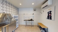 Located at 7425 N Mona Lisa Rd in Tucson, AZ, this purpose-built pet grooming studio is a single-room space designed for efficient, professional pet care. The room is finished with light-toned large-format floor tiles that are both practical and easy to clean. The lower half of the walls features classic white square tiles, providing a hygienic, durable surface, while one accent wall is adorned with a bold geometric patterned tile in black and white, adding a stylish visual focal point to the otherwise clean, functional interior. A stainless steel wash station is positioned along the accent wall, accompanied by a multi-shelf storage unit with transparent doors for organized supply access. A professional grooming table with an adjustable arm and a mounted dryer occupies the center of the room, ready for use. Wall-mounted hooks provide additional storage and organization. A mini-split HVAC unit ensures year-round climate control, while recessed ceiling lighting keeps the space well-lit. A large picture window floods the room with natural light and offers a view of the surrounding Tucson landscape. A glass-panel entry door reinforces the bright, airy atmosphere. Charming pet-themed wall art, including a portrait of a dog named Tucker, adds a welcoming, personable touch to this well-appointed grooming space.