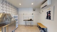 Located at 7425 N Mona Lisa Rd in Tucson, AZ, this purpose-built pet grooming studio is a single-room space designed for efficient, professional pet care. The room is finished with light-toned large-format floor tiles that are both practical and easy to clean. The lower half of the walls features classic white square tiles, providing a hygienic, durable surface, while one accent wall is adorned with a bold geometric patterned tile in black and white, adding a stylish visual focal point to the otherwise clean, functional interior. A stainless steel wash station is positioned along the accent wall, accompanied by a multi-shelf storage unit with transparent doors for organized supply access. A professional grooming table with an adjustable arm and a mounted dryer occupies the center of the room, ready for use. Wall-mounted hooks provide additional storage and organization. A mini-split HVAC unit ensures year-round climate control, while recessed ceiling lighting keeps the space well-lit. A large picture window floods the room with natural light and offers a view of the surrounding Tucson landscape. A glass-panel entry door reinforces the bright, airy atmosphere. Charming pet-themed wall art, including a portrait of a dog named Tucker, adds a welcoming, personable touch to this well-appointed grooming space.