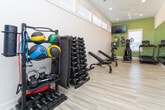 Located at 129 NC-801 S in Bermuda Run, NC, this fitness center is a well-appointed, single-level space designed to support a full range of workout activities. The room features light wood-look flooring that runs throughout, contributing to a clean and airy aesthetic. High ceilings with recessed lighting and a ceiling fan enhance the sense of openness, while a row of clerestory windows along the upper walls floods the space with abundant natural light without sacrificing wall space needed for equipment.

A floor-to-ceiling mirror spans one full wall, visually expanding the room and providing functional feedback during exercise. Multiple flat-screen televisions are mounted at strategic angles, ensuring visibility from various workout stations. The accent walls are painted in a soft sage green, adding a subtle, energizing pop of color against the predominantly white walls and trim.

The fitness center is equipped with treadmills, elliptical trainers, and stationary bikes, several of which feature integrated digital displays. A dedicated free weight area includes an adjustable dumbbell rack and adjustable benches, supporting strength training. Colorful artwork adds a finishing decorative touch, creating an inviting, motivating environment well-suited for a community fitness facility.