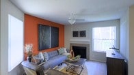 This single-floor living space makes a strong visual impression through a combination of thoughtful design choices and functional features. The living room serves as the centerpiece, anchored by a vibrant burnt-orange accent wall that provides a striking contrast to the neutral gray tones found throughout the rest of the space. A large-scale abstract artwork adorns the accent wall, reinforcing the room's bold aesthetic character.

A tile-surround fireplace adds warmth and a traditional focal point to the room, complemented by a simple white mantel shelf above. Decorative wall art flanks the fireplace, adding visual balance to that corner of the room. A ceiling fan with a light fixture is centrally positioned, providing both comfort and ambient lighting.

Natural light enters through a large window fitted with vertical blinds, brightening the space and creating an airy feel. The flooring appears to be carpeted, contributing to a cozy atmosphere. The room is furnished with a tufted gray sofa, patterned accent pillows, and a geometric metal coffee table atop a layered area rug, completing the contemporary yet welcoming ambiance of the space.