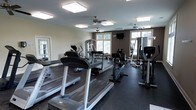 This fitness center offers a comprehensive workout environment designed to accommodate a range of exercise needs. The space is outfitted with multiple treadmills positioned along one side, providing dedicated space for cardio training. Complementing these are upright cycling machines and additional cardio equipment, ensuring variety for aerobic workouts. Toward the back of the room, a multi-station strength training unit is visible alongside weight benches and resistance equipment, supporting full-body strength routines. The flooring consists of dark rubber matting, which provides durability and safety during workouts. Natural light filters into the space through several large windows along the walls, creating a bright and welcoming atmosphere. Overhead, flush-mounted lighting fixtures and dual ceiling fans contribute to consistent illumination and air circulation throughout the room. The walls feature a neutral two-tone color scheme, with lighter upper walls contrasting against a darker lower accent, giving the space a clean and functional aesthetic. A wall-mounted television is also present, offering entertainment during workouts. Overall, the fitness center is thoughtfully arranged to maximize usable floor space while maintaining an open, accessible layout.
