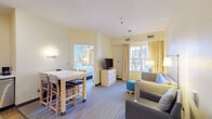 This thoughtfully designed hotel suite offers a comfortable and functional layout spread across a single floor, making it well-suited for extended stays or travelers seeking a home-like environment. The open-plan living and dining area is finished with soft yellow walls and patterned carpet, creating a warm and inviting atmosphere. A high-top dining table with seating anchors one side of the space, while a plush gray sofa, lounge chair, and a teal accent ottoman furnish the living area. Natural light filters in through large windows dressed with gold-toned drapes, adding to the bright, airy feel. A flat-screen TV and dresser unit serve as a media center between the two zones. The fully equipped kitchen features stainless steel appliances — including a full-size refrigerator, dishwasher, and microwave — paired with white cabinetry and tile flooring, offering everything needed for in-suite dining. The separate bedroom provides a serene retreat with a queen bed, light wood furnishings, soft blue accent walls, and generous window views. The bathroom includes a dual-access layout with a dedicated vanity area, a separate bathtub and toilet compartment, and mirrored storage, ensuring privacy and convenience throughout the suite.